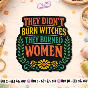 May include: Black embroidered patch with the text "THEY DIDN'T BURN WITCHES THEY BURNED WOMEN" in orange, yellow, and teal. The patch is framed by green leaves and yellow flowers. The patch is a statement piece.