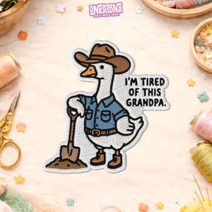 May include: Embroidered patch featuring a white goose wearing a brown cowboy hat, blue shirt, and boots, holding a shovel. The text "I'M TIRED OF THIS GRANDPA." is displayed. The patch has a stitched border and is likely for clothing or accessories.