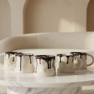 May include: Five off-white ceramic mugs with a distinctive design. Each mug features a textured surface and a dark brown, dripped glaze around the rim, with drips cascading down the sides. The mugs are arranged on a white marble table.