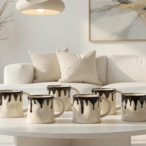 May include: Six cream ceramic mugs with a distinctive design. Each mug has a dark brown, dripped glaze around the rim, creating a stylish contrast. The mugs are arranged on a white table, suggesting a modern home decor theme.