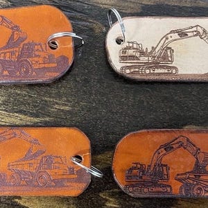 May include: Four oval-shaped leather keychains featuring laser-engraved construction vehicle designs. Two keychains show an excavator loading a dump truck, while the other two depict an excavator digging into a pile of dirt, each with a metal ring.