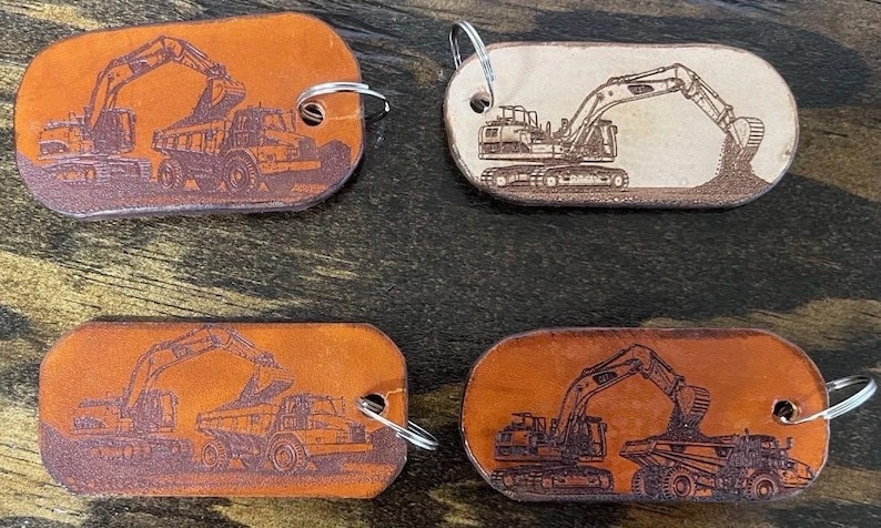 May include: Four oval-shaped leather keychains featuring laser-engraved construction vehicle designs. Two keychains show an excavator loading a dump truck, while the other two depict an excavator digging into a pile of dirt, each with a metal ring.