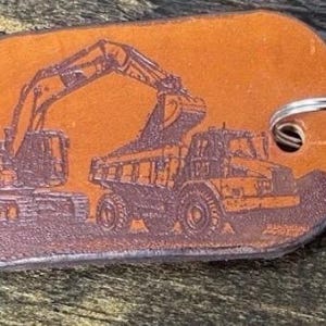 May include: A rectangular tan leather keychain with rounded corners featuring a dark brown laser-engraved illustration of an excavator loading a dump truck. A silver metal split ring is attached through a hole on the right side of the leather tag.