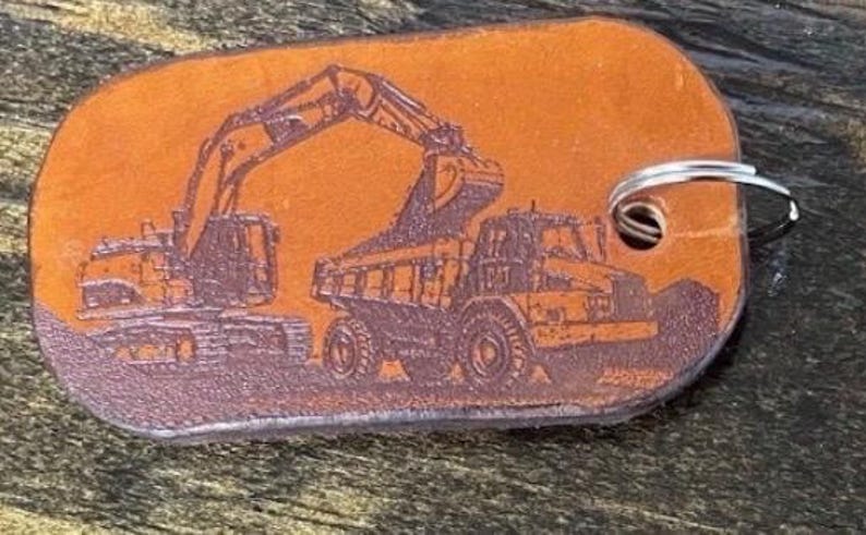 May include: A rectangular tan leather keychain with rounded corners featuring a dark brown laser-engraved illustration of an excavator loading a dump truck. A silver metal split ring is attached through a hole on the right side of the leather tag.