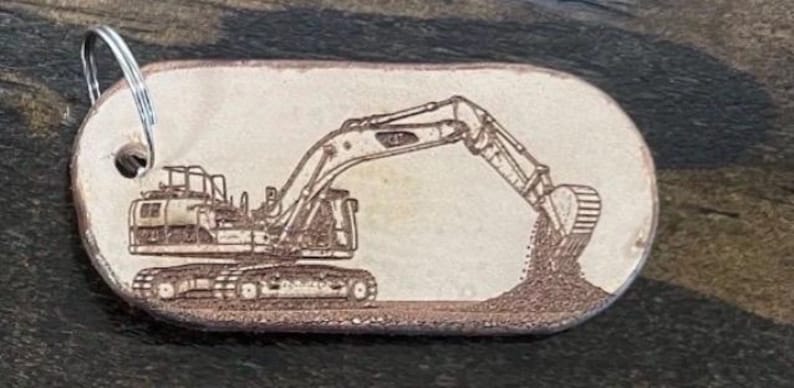 May include: An oval-shaped light tan keychain featuring a detailed laser-engraved illustration of a construction excavator digging into a pile of dirt. The keychain includes a silver-toned metal split ring attached through a hole on the left side.