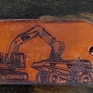 May include: A tan leather keychain with a rounded rectangular shape featuring a dark brown laser-engraved illustration of a tracked excavator loading dirt into the bed of a large dump truck. A silver metal split ring is attached to the right side.