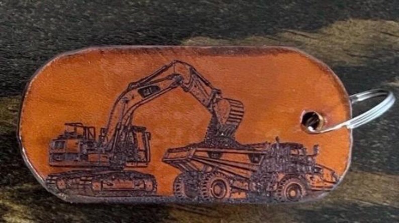 May include: A tan leather keychain with a rounded rectangular shape featuring a dark brown laser-engraved illustration of a tracked excavator loading dirt into the bed of a large dump truck. A silver metal split ring is attached to the right side.