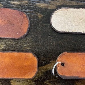 May include: Four rectangular leather tags with rounded corners, each featuring a metal split ring attached through a punched hole. Three tags display varying shades of brown leather, while one tag shows a light beige, textured suede-like finish.