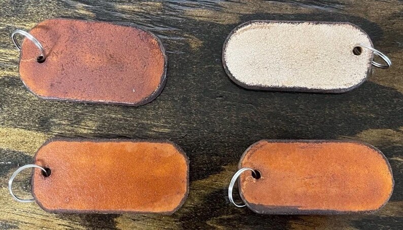May include: Four rectangular leather tags with rounded corners, each featuring a metal split ring attached through a punched hole. Three tags display varying shades of brown leather, while one tag shows a light beige, textured suede-like finish.