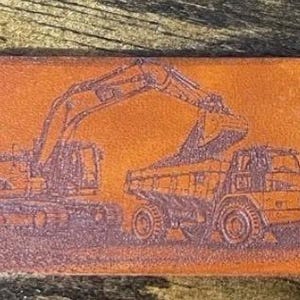 May include: A rectangular brown leather keychain tag featuring a laser-engraved illustration of a Caterpillar excavator loading dirt into a Caterpillar dump truck. The tag has rounded corners and a metal split ring attached through a hole on the right.