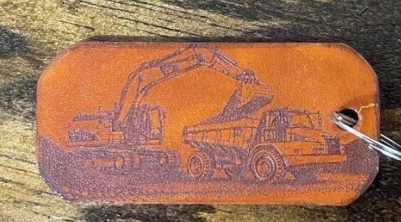 May include: A rectangular brown leather keychain tag featuring a laser-engraved illustration of a Caterpillar excavator loading dirt into a Caterpillar dump truck. The tag has rounded corners and a metal split ring attached through a hole on the right.