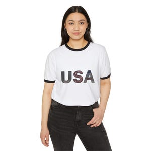 May include: A white ringer t-shirt with black trim on the crew neckline and sleeve cuffs. The front features the text USA in large block letters, with the letter S filled with a red, white, and blue star-patterned graphic design.