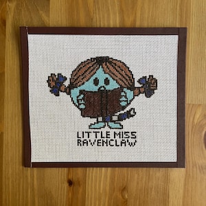 May include: A cross-stitch of a cartoon character with blue skin, brown hair, and a brown dress, holding a book. The text "LITTLE MISS RAVENCLAW" is below the character. The artwork is framed in a dark brown border.