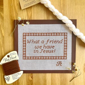 May include: A cross-stitch project with the text "What a friend we have in Jesus!" in brown thread. The project is on a white canvas with a brown border. A spool of brown thread and a length of white yarn are also present.
