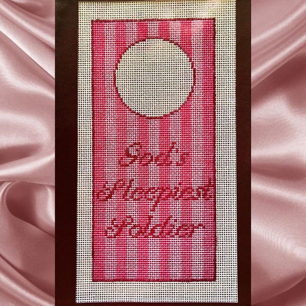Può includere: Una tela rettangolare per ricamo con un motivo a righe verticali rosa e bianche. Il design include un ritaglio circolare nella parte superiore e il testo ricamato "God's sleepiest soldier" in un carattere corsivo rosa scuro al centro.