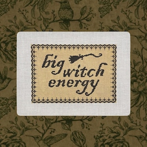May include: A rectangular needlepoint canvas featuring the black stitched text "big witch energy" with a small broomstick icon above the word witch, all enclosed within a decorative black and tan geometric border on a tan background.