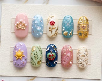 May include: A set of ten artificial nails, each uniquely designed with various themes. Designs include a gold crown, a glass slipper, a rose, a mermaid tail, a heart, a sun, leaves, an apple, feathers, and script. The nails are arranged on a white card.