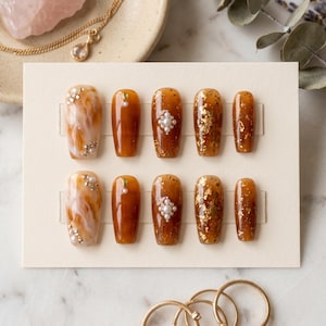 May include: A set of ten press-on nails in various designs. Some nails feature a marbled effect, others are solid brown, and some have gold leaf or pearl accents. The nails are arranged on a cream-colored card.