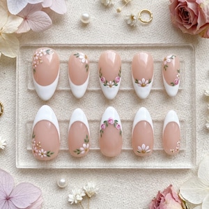 Pode incluir: Um conjunto de dez unhas artificiais com base nude, pontas brancas e desenhos florais. As unhas apresentam vários padrões florais, incluindo pequenas flores brancas, rosas cor-de-rosa e detalhes dourados. Dispostas numa tela de plástico transparente.