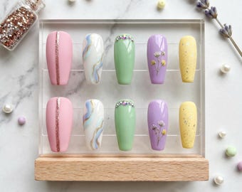 Puede incluir: Una exhibición de diez uñas postizas en varios diseños y colores. Las uñas están dispuestas en un soporte acrílico transparente con una base de madera. Los colores incluyen rosa, blanco, verde, morado y amarillo. Algunas uñas presentan purpurina, efectos de mármol o detalles florales.