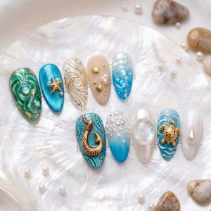May include: Ten artificial nails with a marine theme. The nails are decorated with teal, white, and gold designs, including starfish, seashells, and pearl embellishments. The nails are displayed on a white, textured surface, evoking a seaside aesthetic.