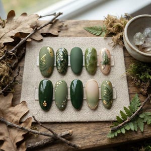Puede incluir: Un juego de diez uñas postizas ovaladas con varios tonos de verde, incluyendo oliva y verde bosque. Los diseños incluyen detalles de pan de oro, motivos de helechos y hojas pintados a mano, acabado mate y adornos dorados.