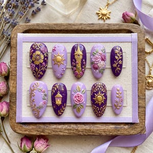 May include: A collection of ten purple and lavender press-on nails with detailed designs. Each nail showcases gold accents, such as floral patterns, sunbursts, and a tower. The nails are presented on a wooden tray with lavender ribbon and dried flowers.