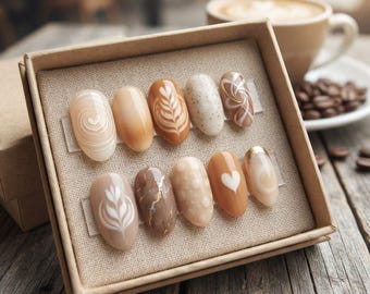 Puede incluir: Un juego de diez uñas postizas ovaladas dispuestas en una caja de cartón. Las uñas presentan varios diseños inspirados en el café, incluyendo arte latte, patrones de mármol, lunares, un corazón blanco y detalles dorados en tonos beige y marrón.