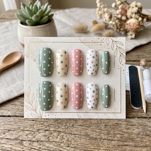 May include: A set of ten press-on nails arranged on a rectangular display card. The nails feature a glossy finish with polka dot patterns in shades of sage green, cream, and dusty rose. The display card includes a delicate gold leaf line art.