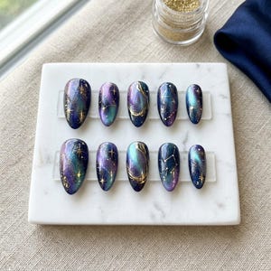 May include: A set of ten almond-shaped press-on nails featuring a deep galaxy-inspired design with swirling shades of dark blue, purple, and teal. The nails are decorated with metallic gold hand-painted stars, constellations, and fine glitter accents.