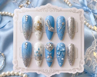 Puede incluir: Un juego de diez uñas postizas en forma de almendra puntiaguda, con una paleta de colores azul claro y blanco. Los diseños incluyen degradados de purpurina plateada, un carruaje dorado, un zapato, una varita mágica, un castillo y un lazo.