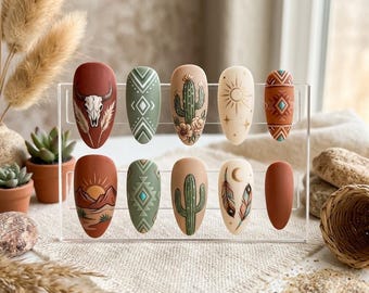 Puede incluir: Un juego de diez uñas postizas en forma de almendra sobre un soporte de acrílico transparente. Los diseños presentan tonos tierra mate, incluyendo terracota, verde salvia y crema, con ilustraciones de un cráneo de toro, cactus, soles y plumas.