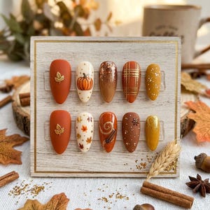 Könnte beinhalten: Ein Set aus zehn mandelförmigen Press-on-Nägeln auf einem Holzbrett. Die Designs zeigen herbstliche Motive wie mattes Orange mit Blattgold, Creme mit Kürbissen und Blättern, braunen Glitzer, orangefarbenes Karomuster, Senfgelb und Zimtstangen.