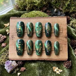 Peut inclure: Un ensemble de dix faux ongles vert émeraude avec divers motifs. Certains ongles présentent des croissants de lune et des étoiles dorées, d'autres des motifs de feuilles d'or et des motifs géométriques dorés. Les ongles sont présentés sur une planche de bois.