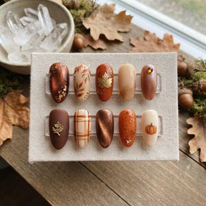 Op de afbeelding: Een set van tien amandelvormige press-on nagels met herfstthema in tinten bruin, oranje en crème. De nagels zijn voorzien van metallic afwerkingen, bladgoudaccenten, een ruitpatroon, een bladgoudbedel en een klein 3D-pompoendetail.