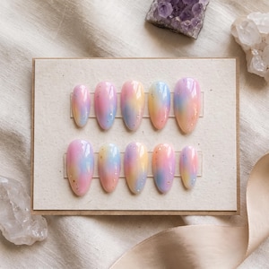 Puede incluir: Un juego de diez uñas postizas en forma de almendra dispuestas en dos filas sobre una tarjeta rectangular. Cada uña presenta un degradado suave de colores pastel rosa, amarillo y azul, acentuado con pequeñas láminas de oro dispersas.