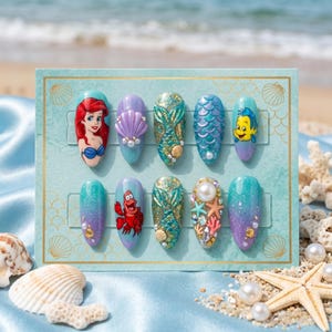 May include: A set of ten artificial nails with a mermaid theme. Each nail features a different design, including Ariel, Flounder, and Sebastian, along with mermaid tails, shells, and pearls. The nails are displayed on a blue card with a seashell border.
