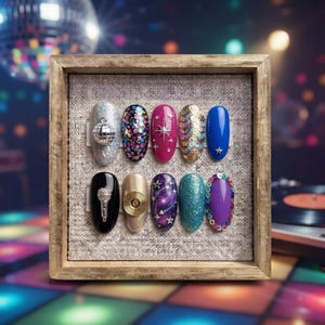 Könnte beinhalten: Ein Set aus zehn künstlichen Fingernägeln in einem Holzrahmen mit verschiedenen Designs, darunter ein silberner Discokugel-Anhänger, bunte Pailletten, ein Mikrofon-Anhänger, eine goldene Schallplatte, Sternmotive, Galaxienmuster und Glitzer.