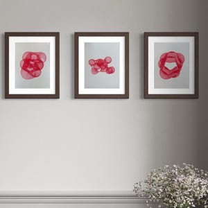 May include: Three framed wall art prints featuring abstract red circular line patterns on a white background, displayed in dark brown frames. The artwork is arranged in a horizontal row on a light gray wall above a black chair and a marble pedestal.