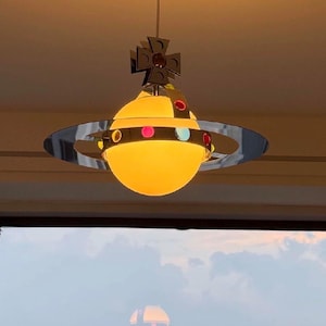 Può includere: Una lampada a sospensione sferica caratterizzata da un globo luminoso giallo caldo circondato da un anello color argento lucido decorato con piccoli accenti circolari colorati. Un ornamento a forma di croce argentata sovrasta la sfera.