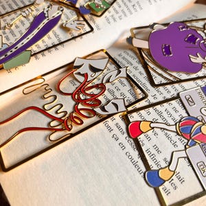 May include: A collection of gold-toned metal bookmarks featuring various enamel designs, including a theatre mask motif with red coiled wire, a purple character, a colourful star-patterned piece, and a computer error window graphic.