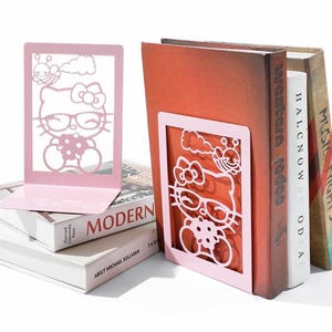 May include: Two light pink metal bookends featuring a laser-cut design of a cartoon cat wearing glasses and holding a bouquet of flowers, with a small bee and cloud above. The bookends are shown supporting a small stack of hardback books.