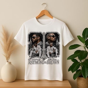 May include: A white short-sleeve t-shirt featuring a graphic print of two men in suits, identified as Usher and Chris Brown, with the text "Usher Raymond & Chris Brown" displayed in a decorative, glittery, black and silver frame design.