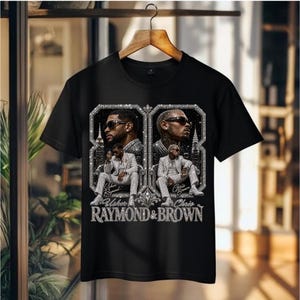 May include: A black short-sleeve t-shirt featuring a graphic print of Usher and Chris Brown. The design includes two framed portraits of the artists in suits, set against a city skyline background with the text Usher Raymond & Brown below.