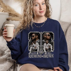 May include: A navy blue crewneck sweatshirt featuring a graphic print of Usher and Chris Brown. The design includes two framed portraits of the artists in suits, with the text "Usher Raymond" and "Chris Brown" below them and "RAYMOND & BROWN" at the base.