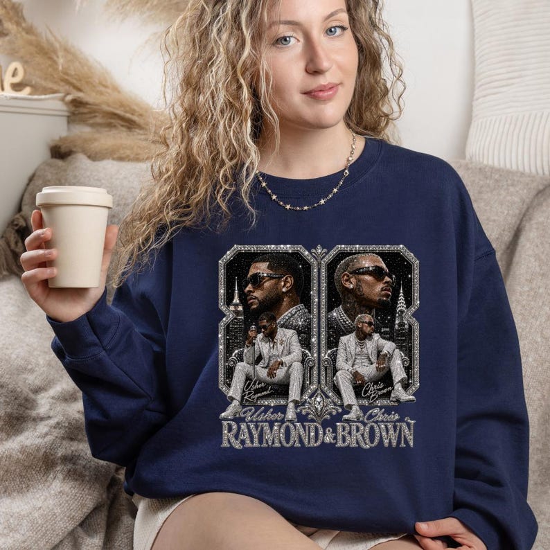May include: A navy blue crewneck sweatshirt featuring a graphic print of Usher and Chris Brown. The design includes two framed portraits of the artists in suits, with the text "Usher Raymond" and "Chris Brown" below them and "RAYMOND & BROWN" at the base.