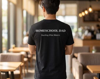 Op de afbeelding: Zwart t-shirt met de witte tekst "HOMESCHOOL DAD" gedrukt op de bovenrug, met "Teaching What Matters" eronder. Het t-shirt wordt gedragen door een persoon in een café.