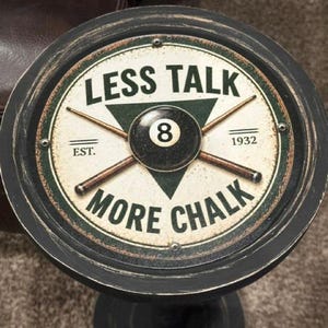 May include: A round, black metal wall sign featuring a cream-colored center with a green triangle, two crossed pool cues, and a black 8-ball. The text reads LESS TALK, MORE CHALK, and EST. 1932. The sign has a distressed finish and four visible screws.