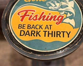 May include: A round decorative sign with a black frame featuring a graphic of a fish and the text Gone Fishing BE BACK AT DARK THIRTY. The background is divided into teal and yellow sections with red lettering for the word Fishing.