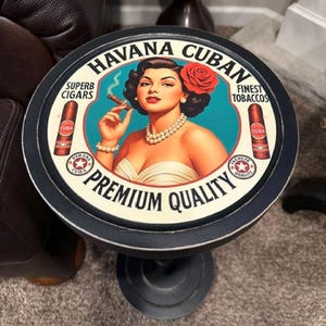 May include: A round black side table featuring a vintage-style graphic on the top. The design includes the text "HAVANA CUBAN PREMIUM QUALITY," "SUPERB CIGARS," and "FINEST TOBACCOS," with an illustration of a person holding a cigar.